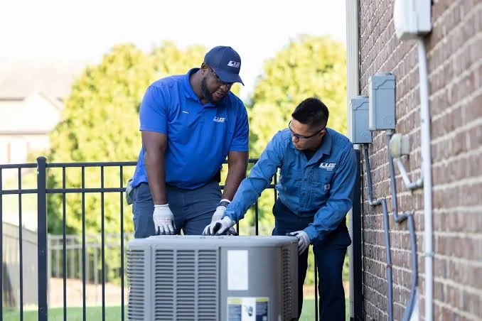 Two technicians working on an outdoor condenser beside a brick building
